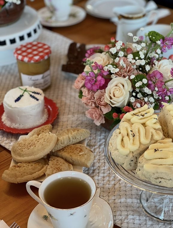 An afternoon tea table with a lavender-decorated mini cake, crumpets, scones topped with lemon crème, fresh flowers and a cup of tea