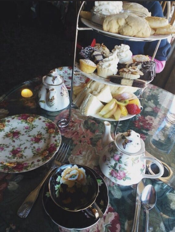 A three-tier afternoon tea stand with scones and cream, petite sweets, tea sandwiches and fruit, beside floral china teapots and a black floral teacup