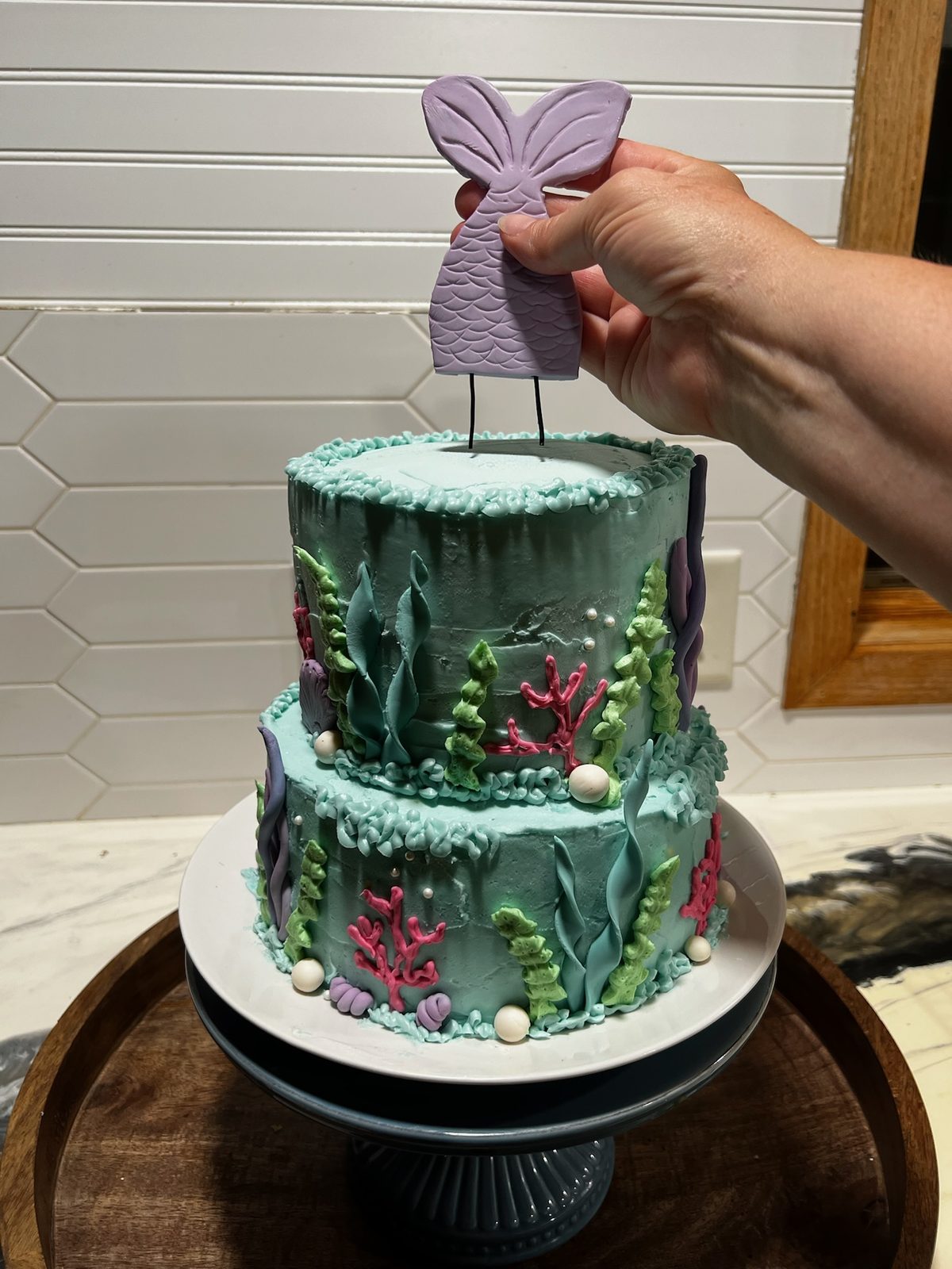 A two-tier teal mermaid cake with fondant seaweed, coral and pearls, with a lavender mermaid-tail topper