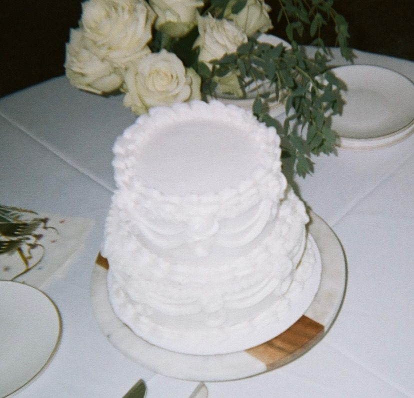 A two-tier white wedding cake with piped buttercream frills beside white roses