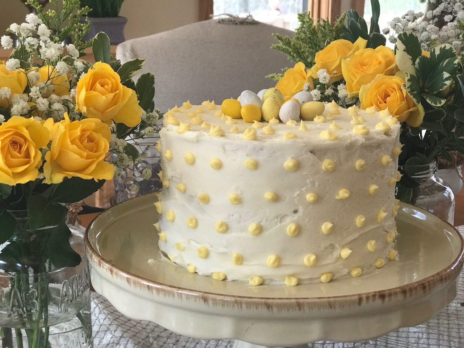 A white layer cake with yellow buttercream dots and candy eggs, between vases of yellow roses