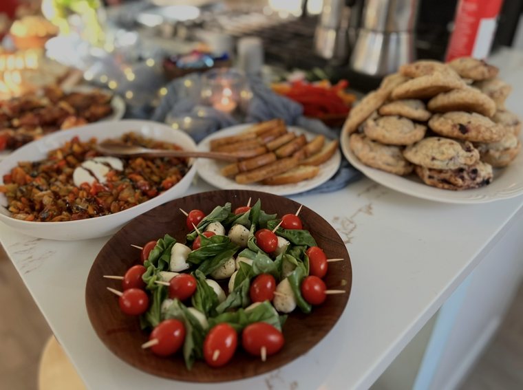 A wooden bowl of caprese skewers with cherry tomatoes, fresh mozzarella and basil, with cookies and appetizers behind