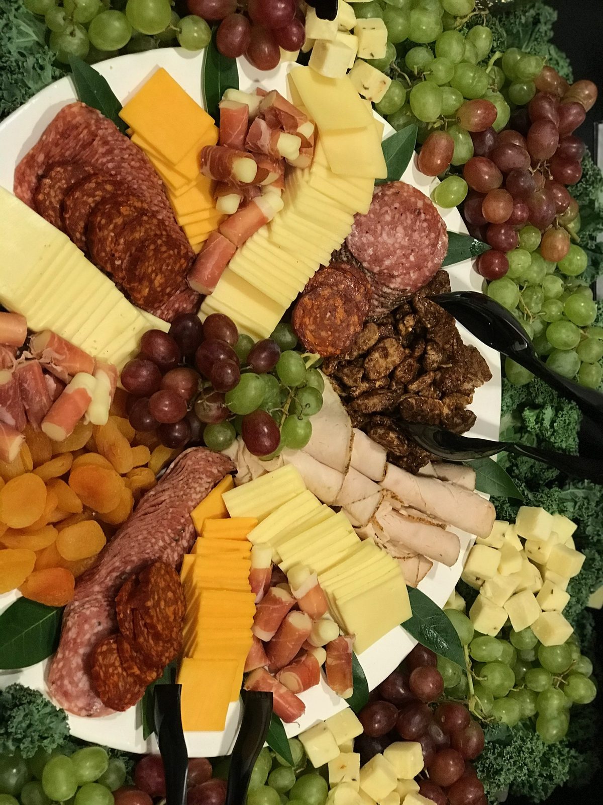 A catering platter of salami, prosciutto-wrapped cheese, sliced cheeses, candied pecans, dried apricots and grapes on a bed of kale