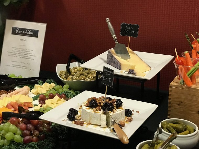 A catering table with a wedge of aged Gouda, a wheel of brie topped with blackberries, candied pecans and honey, marinated olives, vegetable cups and a printed event menu