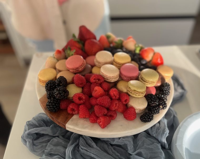 A round marble and wood board piled with French macarons, strawberries, raspberries, blackberries and blueberries