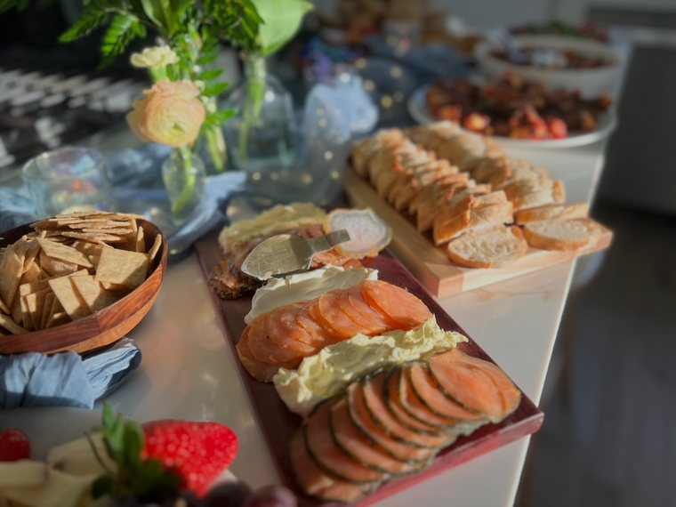 A board of sliced smoked salmon with herbed butter and cream cheese, beside crackers and sliced baguette